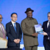 Four men in formal attire on a stage; one in a khaki uniform hands a booklet to another man in a suit during an awards ceremony.