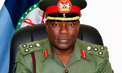 Front-facing high-ranking military officer in green dress uniform with medals, seated in a chair with a flag behind.