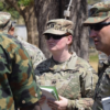 Female U.S. Army soldier in camouflage with sunglasses holds papers during briefing with fellow soldiers outdoors orients toward instructions on a field activity.