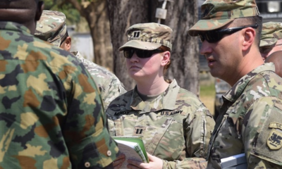 Female U.S. Army soldier in camouflage with sunglasses holds papers during briefing with fellow soldiers outdoors orients toward instructions on a field activity.
