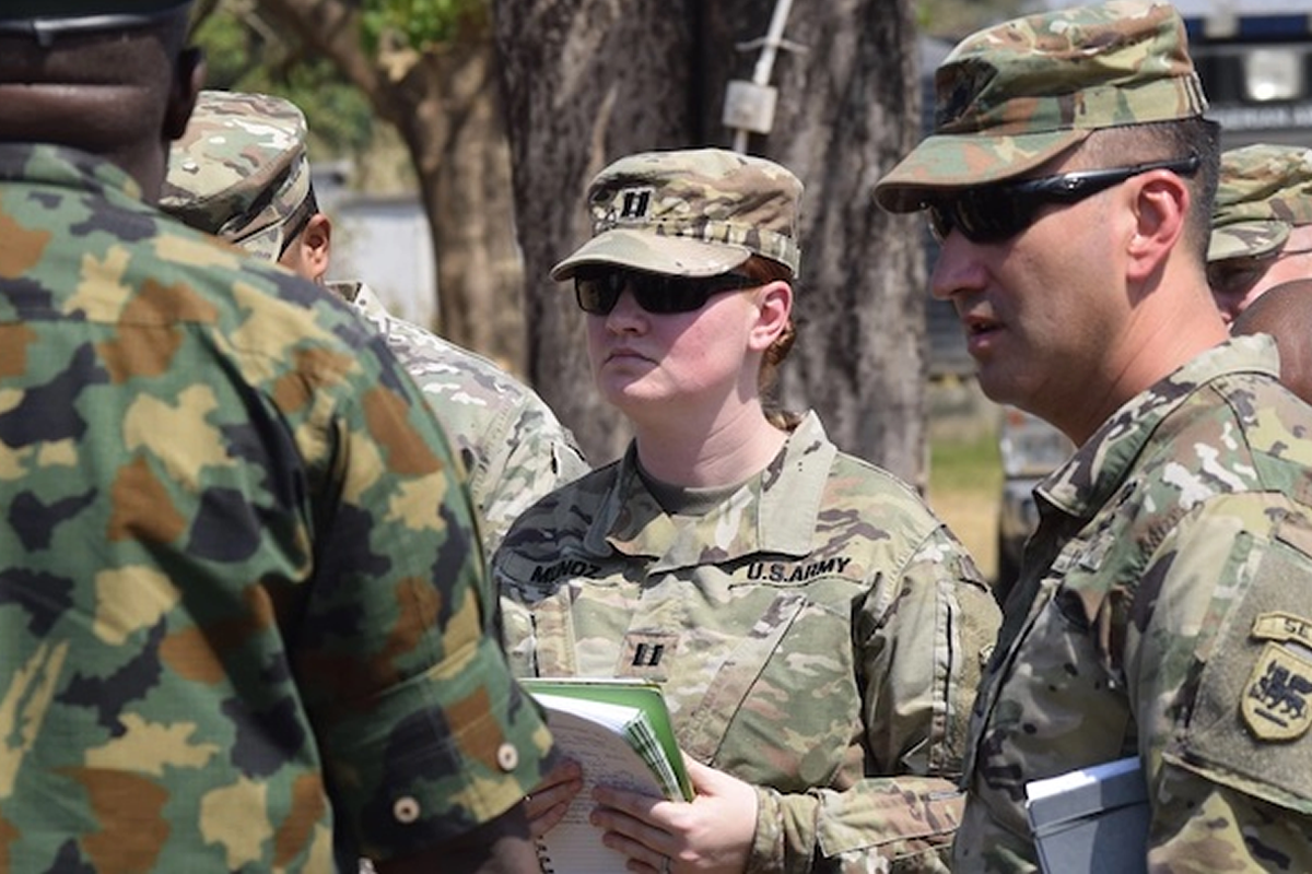 Female U.S. Army soldier in camouflage with sunglasses holds papers during briefing with fellow soldiers outdoors orients toward instructions on a field activity.