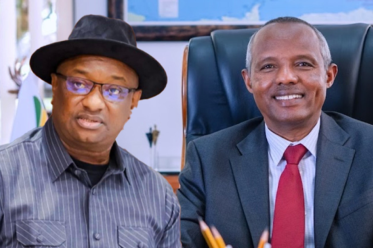 Two men in an office pose for a photo: one wearing a black hat and striped shirt, the other in a gray suit with a red tie, both smiling.
