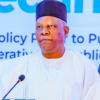 Man in white traditional attire and black cap speaks at a podium with a microphone.