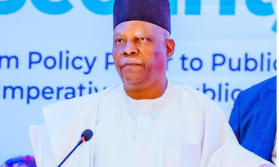 Man in white traditional attire and black cap speaks at a podium with a microphone.