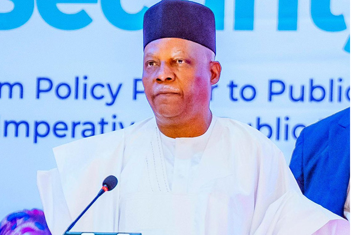 Man in white traditional attire and black cap speaks at a podium with a microphone.