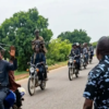 Group of men on motorcycles on a rural road; one rider raises a hand as others carry rifles.