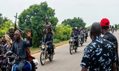 Group of men on motorcycles on a rural road; one rider raises a hand as others carry rifles.