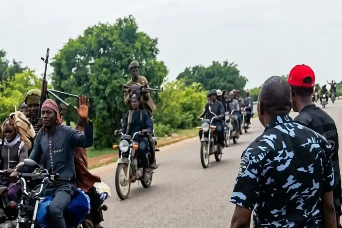 Group of men on motorcycles on a rural road; one rider raises a hand as others carry rifles.