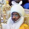 Man wearing a white ceremonial headwrap with ear-like flaps and glasses, seated on a gold, ornate chair in a formal setting with a yellow patterned robe.