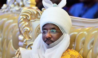 Man wearing a white ceremonial headwrap with ear-like flaps and glasses, seated on a gold, ornate chair in a formal setting with a yellow patterned robe.