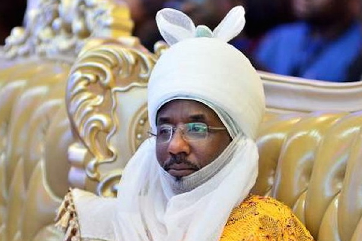 Man wearing a white ceremonial headwrap with ear-like flaps and glasses, seated on a gold, ornate chair in a formal setting with a yellow patterned robe.