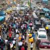 Crowd of protesters marching down a busy city street, filling the road with vehicles on both sides awaiting passage.