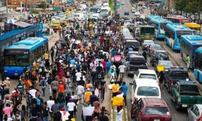 Crowd of protesters marching down a busy city street, filling the road with vehicles on both sides awaiting passage.