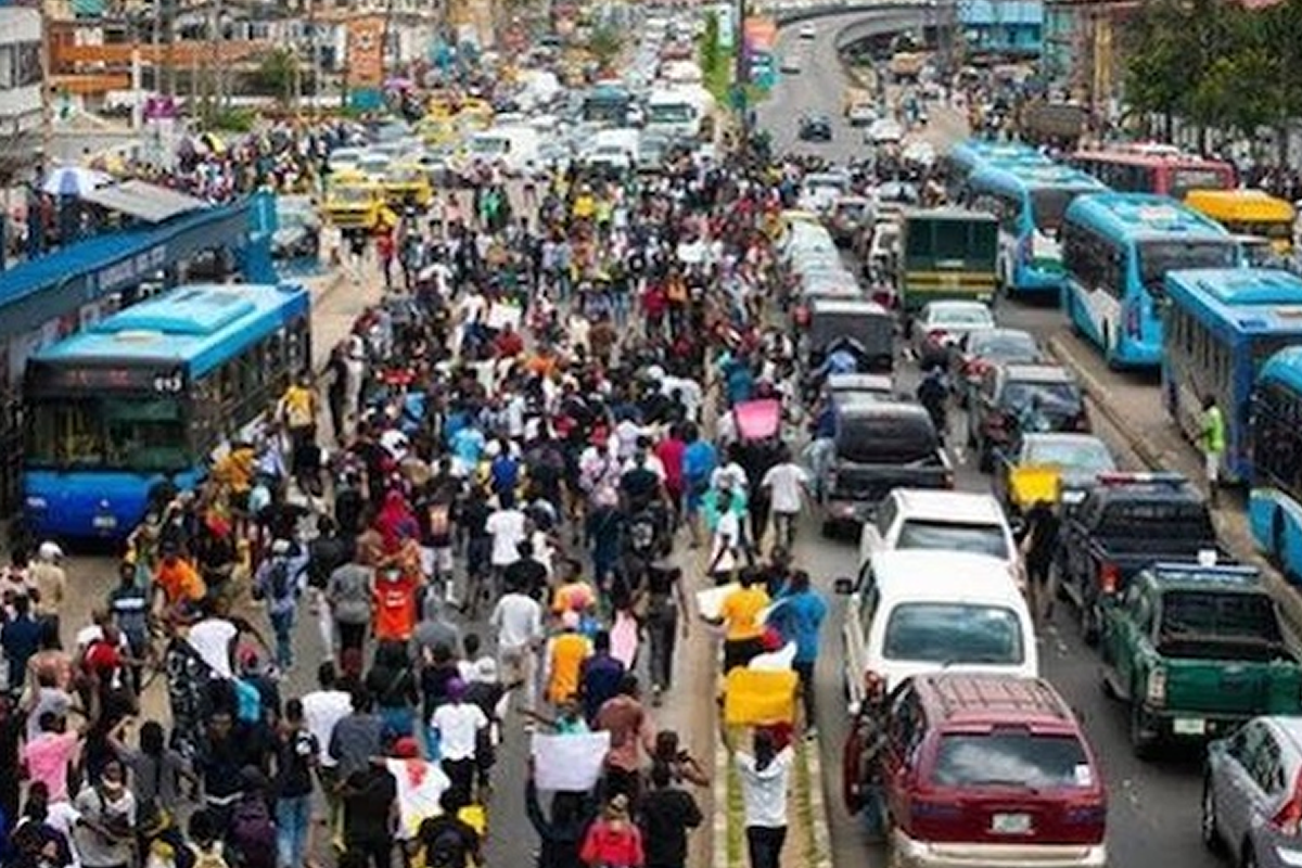 Crowd of protesters marching down a busy city street, filling the road with vehicles on both sides awaiting passage.