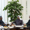 Four men in traditional Arab attire sit and chat around a coffee table with fruit bowls in a bright indoor lounge with a large plant behind them.
