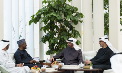 Four men in traditional Arab attire sit and chat around a coffee table with fruit bowls in a bright indoor lounge with a large plant behind them.