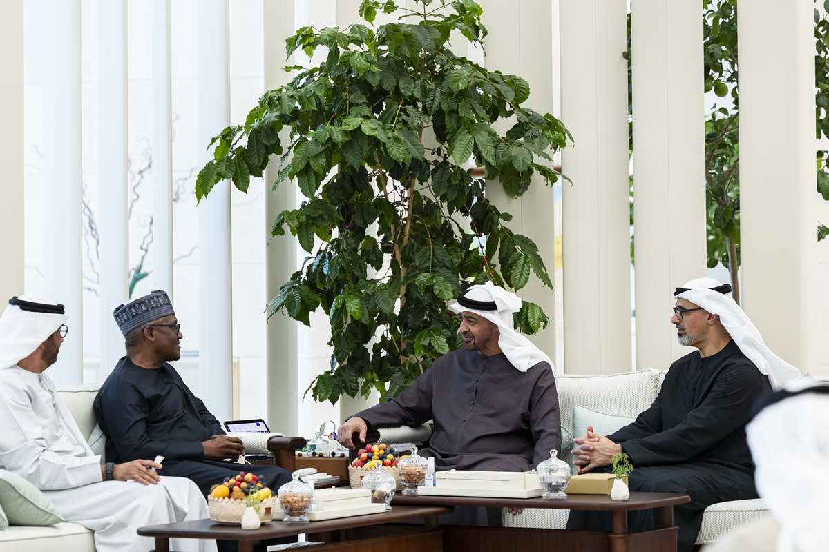 Four men in traditional Arab attire sit and chat around a coffee table with fruit bowls in a bright indoor lounge with a large plant behind them.