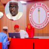 Senate chamber scene with the Seal of the Senate of Nigeria on the wall and three officials at a red desk discussing over a laptop.