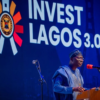 Man in blue traditional attire speaks at a podium during the 'Invest Lagos 3.0' event; Lagos state seal on the lectern and a large banner behind him.