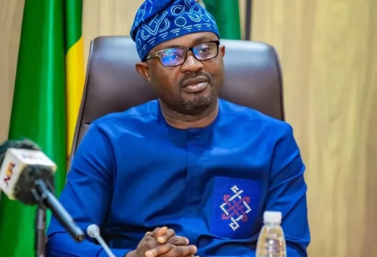 Man in blue traditional outfit and glasses sits at a desk with a microphone in front, hands clasped, against a backdrop of green and yellow flags.