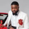 Man in a white suit with a black bow tie leaning on a red convertible, with red flowers nearby.