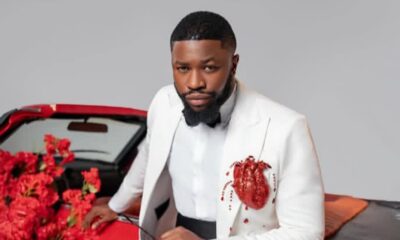Man in a white suit with a black bow tie leaning on a red convertible, with red flowers nearby.