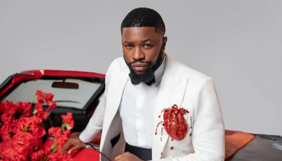 Man in a white suit with a black bow tie leaning on a red convertible, with red flowers nearby.