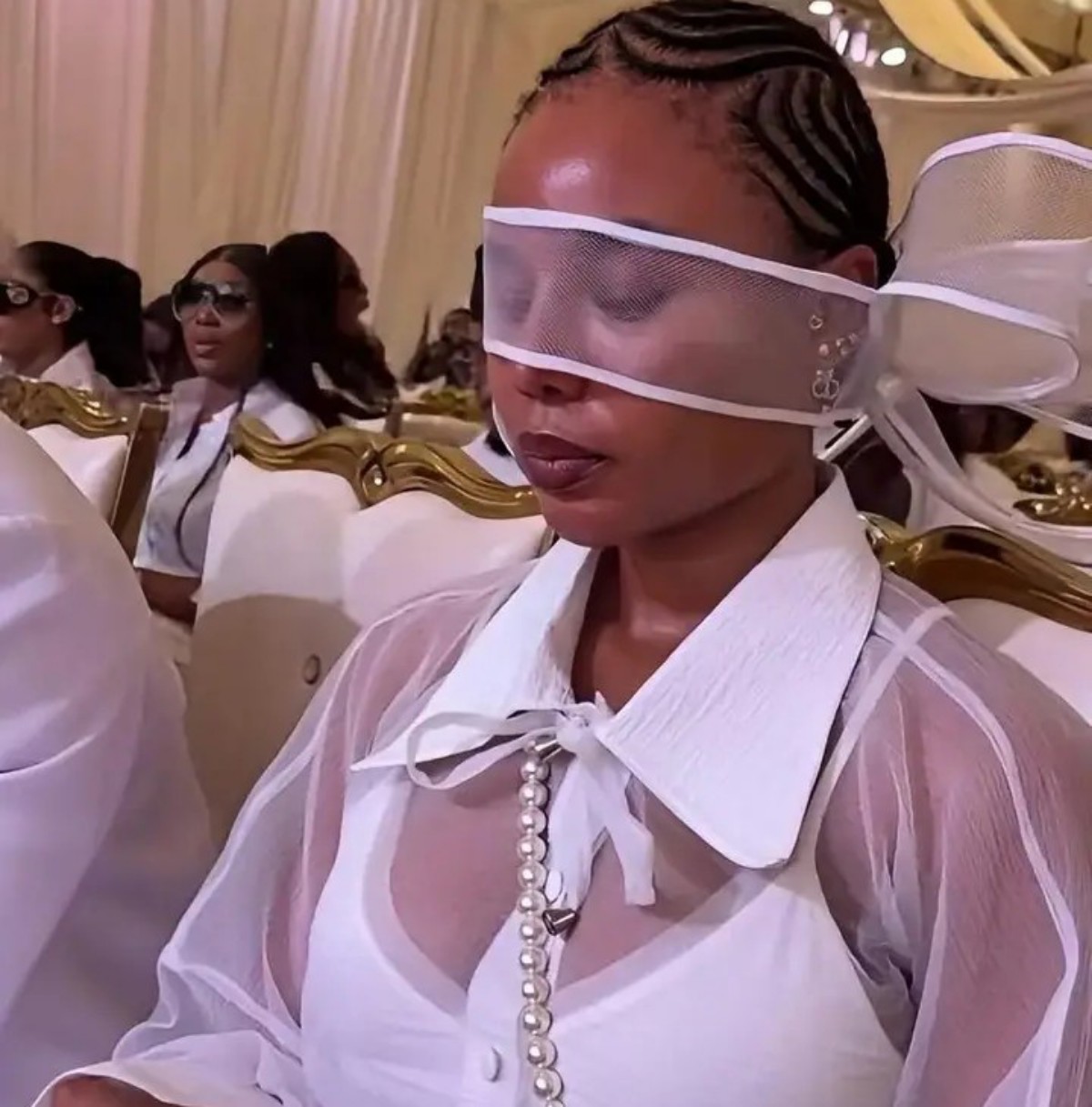 Woman at a formal event wearing a translucent white visor over her eyes and a sheer white outfit with a pearl necklace.