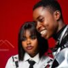 Two young people pose together against a red backdrop; woman wears a white shirt and tie, man in a shiny jacket, AFROKONNECT logo visible in the background.