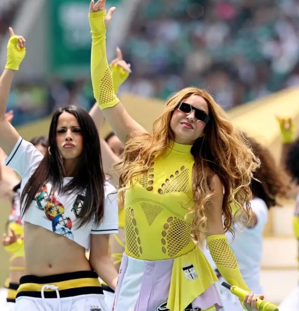 Two women dancing at a parade, one in a neon yellow mesh outfit with gloves and sunglasses, raising her arm with others nearby.
