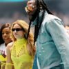 Tall man with braided hair and orange sunglasses in a light blue denim jacket beside two women in neon yellow outfits at an event.