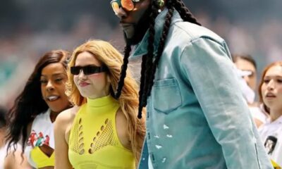Tall man with braided hair and orange sunglasses in a light blue denim jacket beside two women in neon yellow outfits at an event.