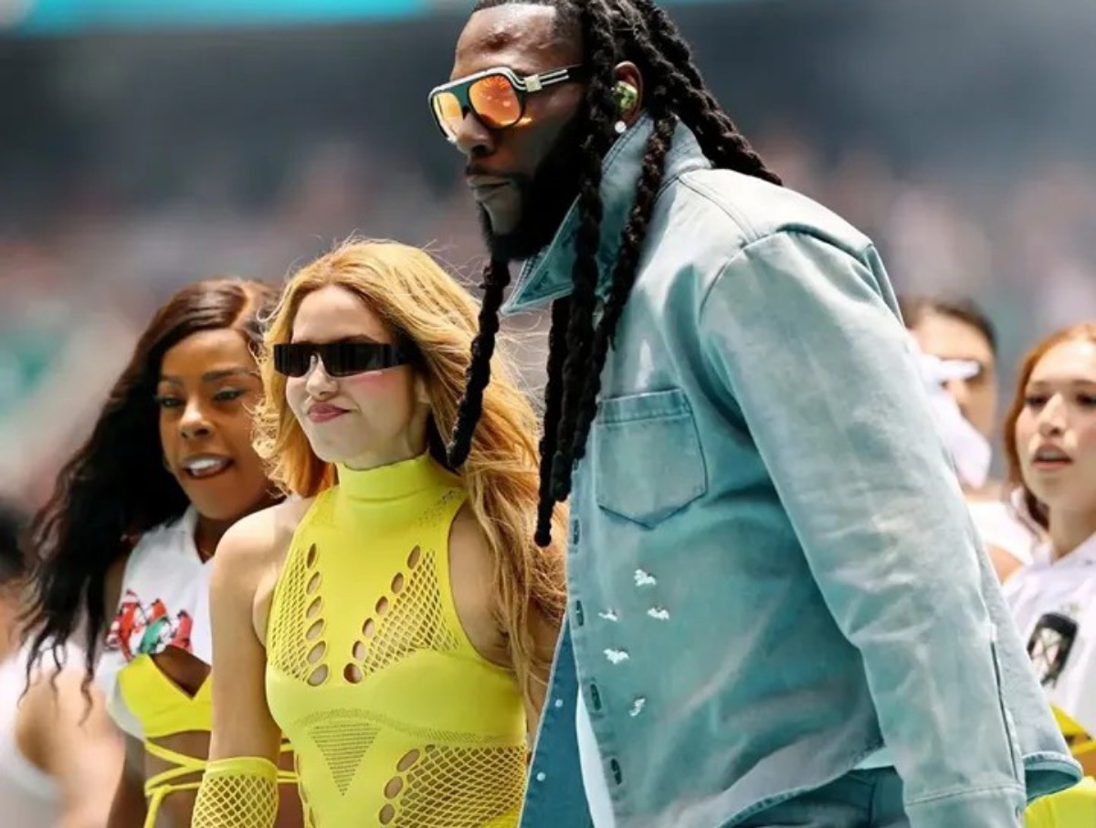 Tall man with braided hair and orange sunglasses in a light blue denim jacket beside two women in neon yellow outfits at an event.
