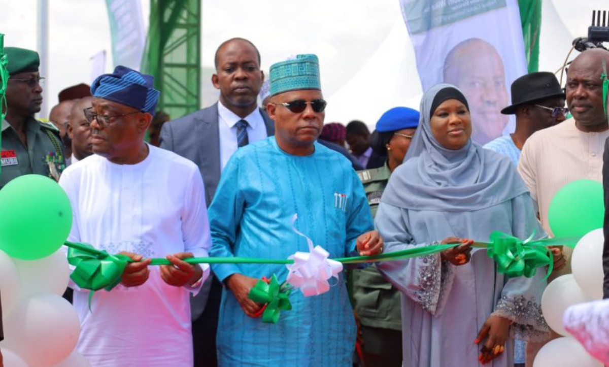 Dignitaries cut a green ribbon at a ceremonial event, with balloons and a banner in the background.