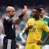 Soccer referee in black shows a red card to a South Africa player in a yellow jersey.