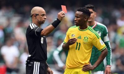 Soccer referee in black shows a red card to a South Africa player in a yellow jersey.