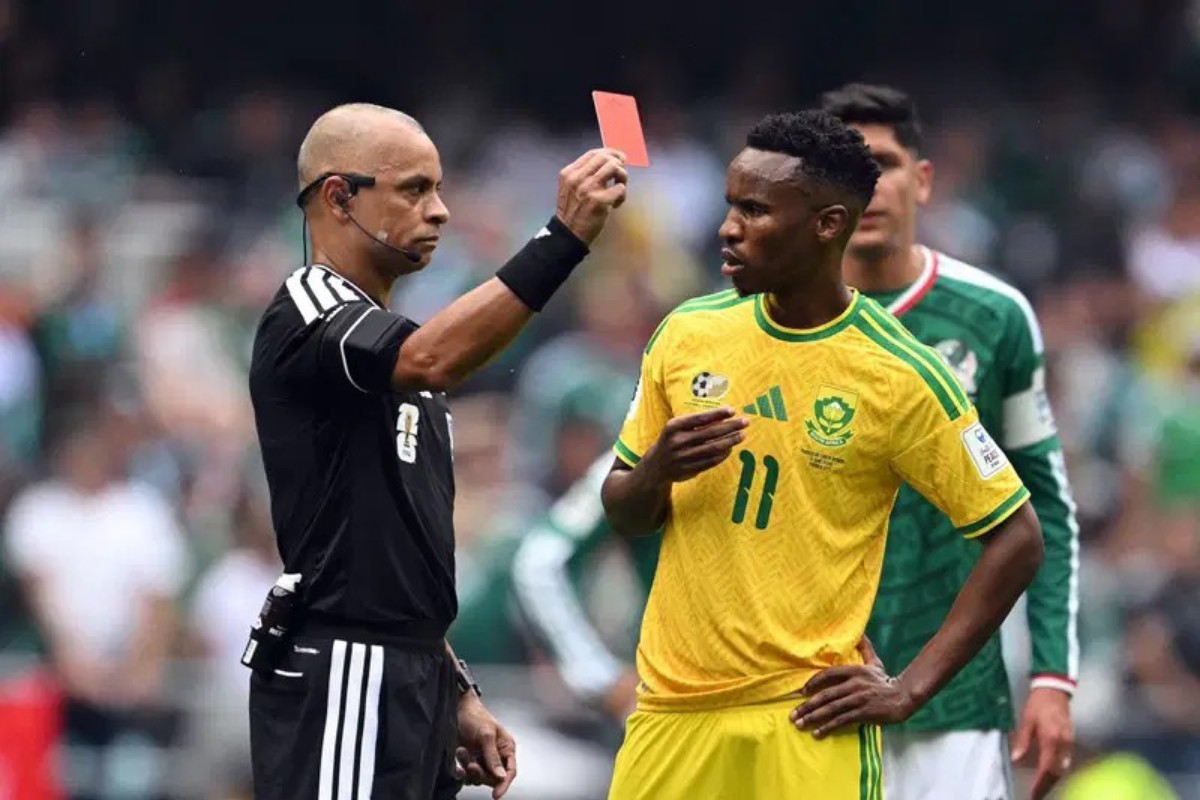 Soccer referee in black shows a red card to a South Africa player in a yellow jersey.