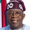 Smiling man in traditional blue attire and maroon headwrap with round glasses, flags in the background.