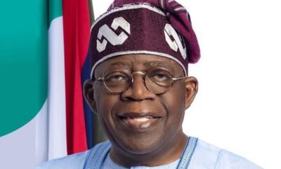 Smiling man in traditional blue attire and maroon headwrap with round glasses, flags in the background.