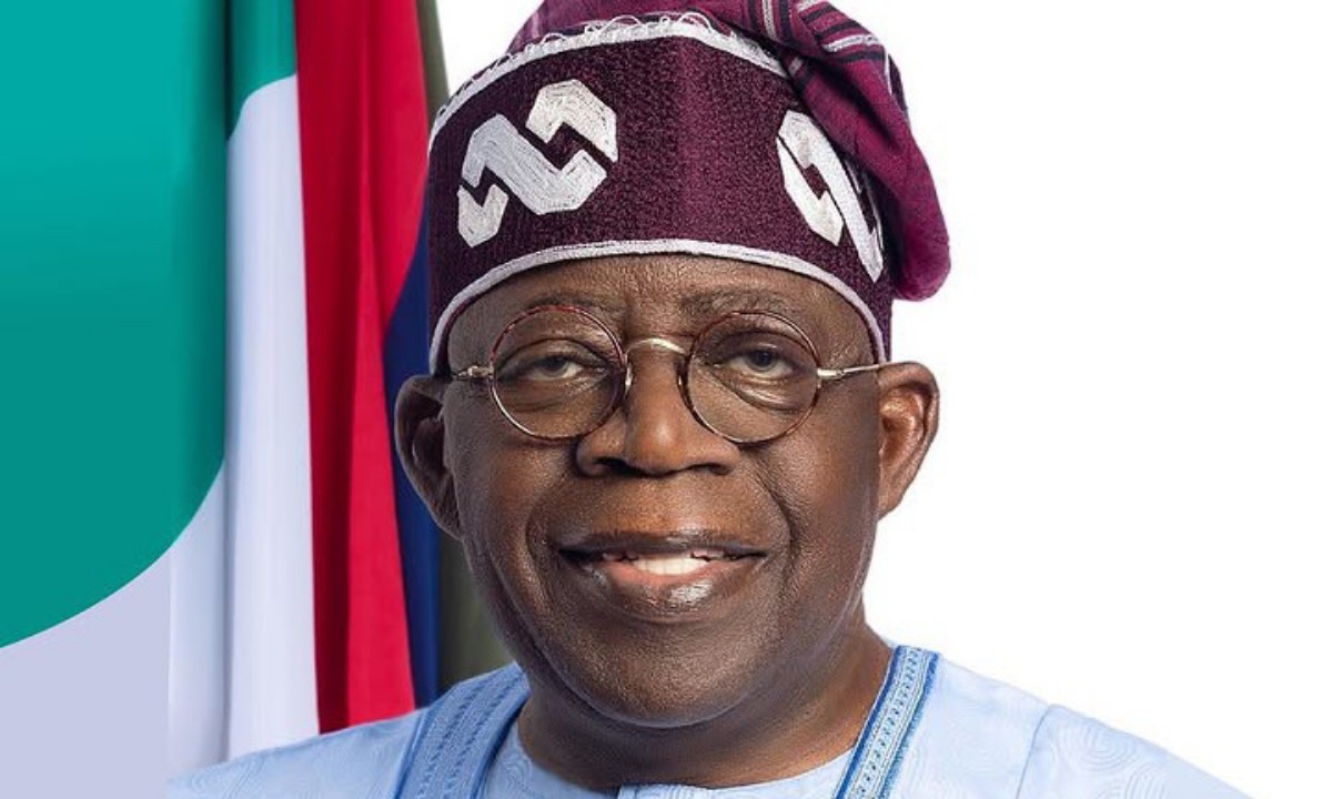 Smiling man in traditional blue attire and maroon headwrap with round glasses, flags in the background.