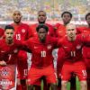 Canada national soccer team poses for a group photo on the field in red uniforms, stadium full of fans in the background, banner reading CANADA held by player in front.