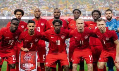 Canada national soccer team poses for a group photo on the field in red uniforms, stadium full of fans in the background, banner reading CANADA held by player in front.