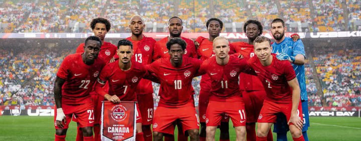 Canada national soccer team poses for a group photo on the field in red uniforms, stadium full of fans in the background, banner reading CANADA held by player in front.