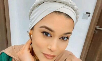 Portrait of a woman wearing a light gray braided headwrap and beige hijab, resting her face on her hand in a neutral pose indoors.
