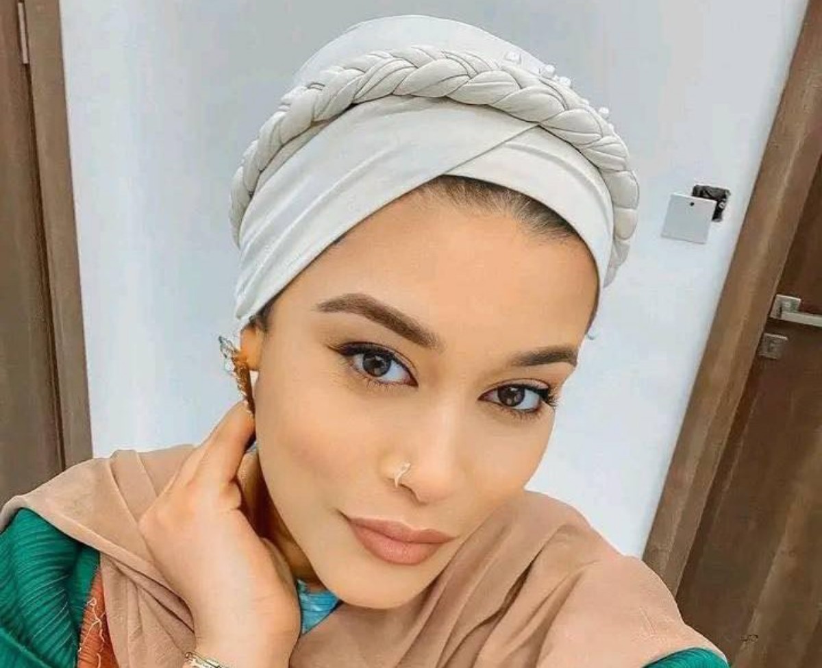 Portrait of a woman wearing a light gray braided headwrap and beige hijab, resting her face on her hand in a neutral pose indoors.