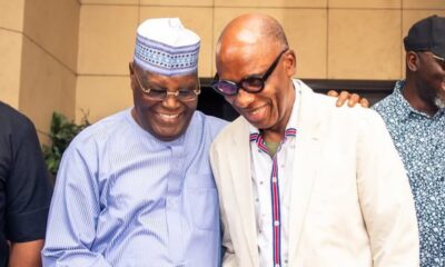 Two men smile and lean together outdoors; one wears a traditional cap and striped shirt, the other a light blazer and colorful shirt, with a third man nearby.