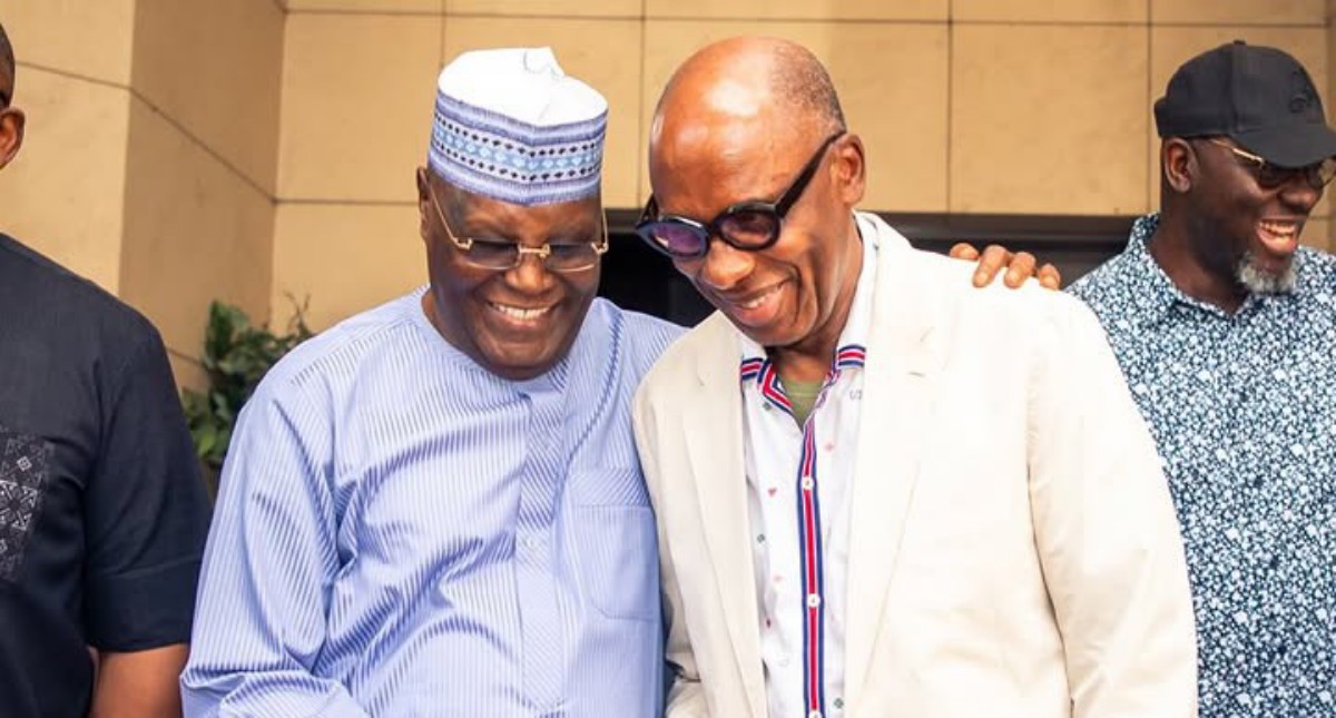 Two men smile and lean together outdoors; one wears a traditional cap and striped shirt, the other a light blazer and colorful shirt, with a third man nearby.