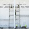 Construction workers in safety vests assemble scaffolding against a white memorial wall with large engraved text up front.