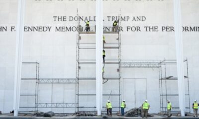 Construction workers in safety vests assemble scaffolding against a white memorial wall with large engraved text up front.