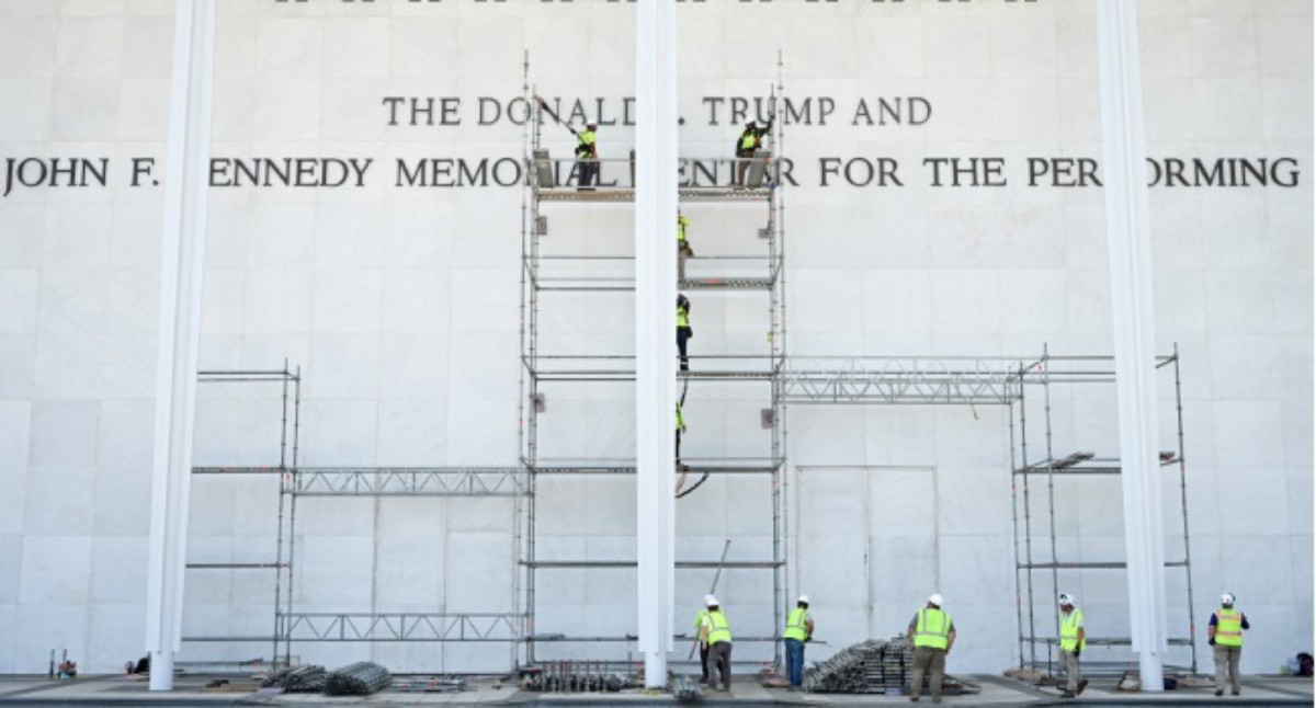Construction workers in safety vests assemble scaffolding against a white memorial wall with large engraved text up front.